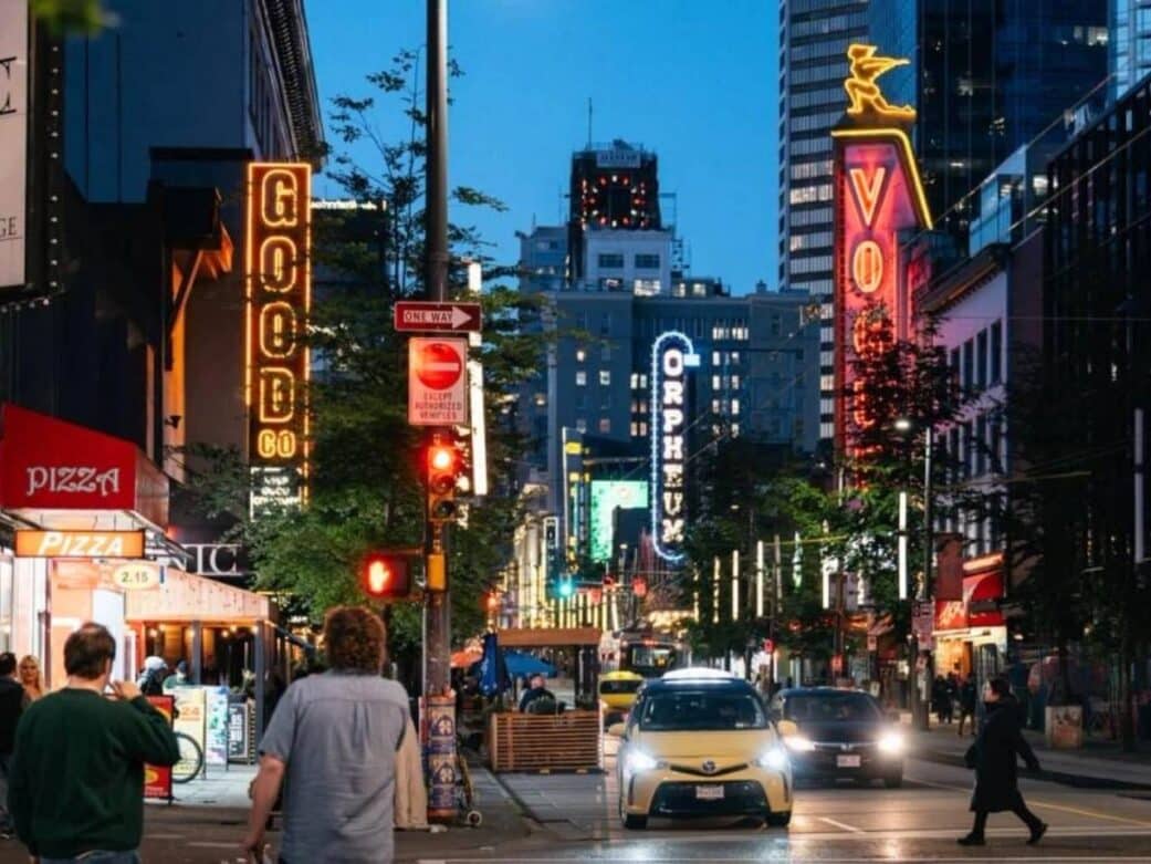 Busy Granville city street at dusk, vibrant neon signs including "GOOD CO" and "ORPHEUM," with cars and people crossing. Energetic urban atmosphere.