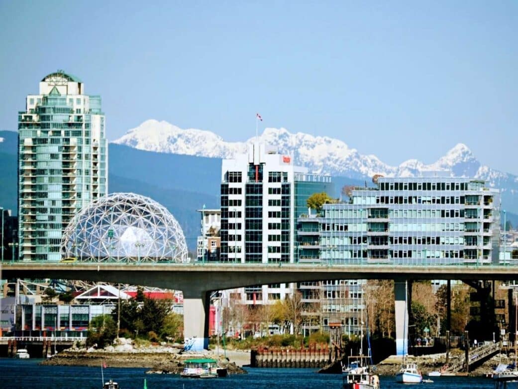 Cityscape featuring the geodesic dome of science world and modern buildings with glass facades, set against snow-capped mountains and a blue sky. Boats float in the foreground of olympic village in Vancouver