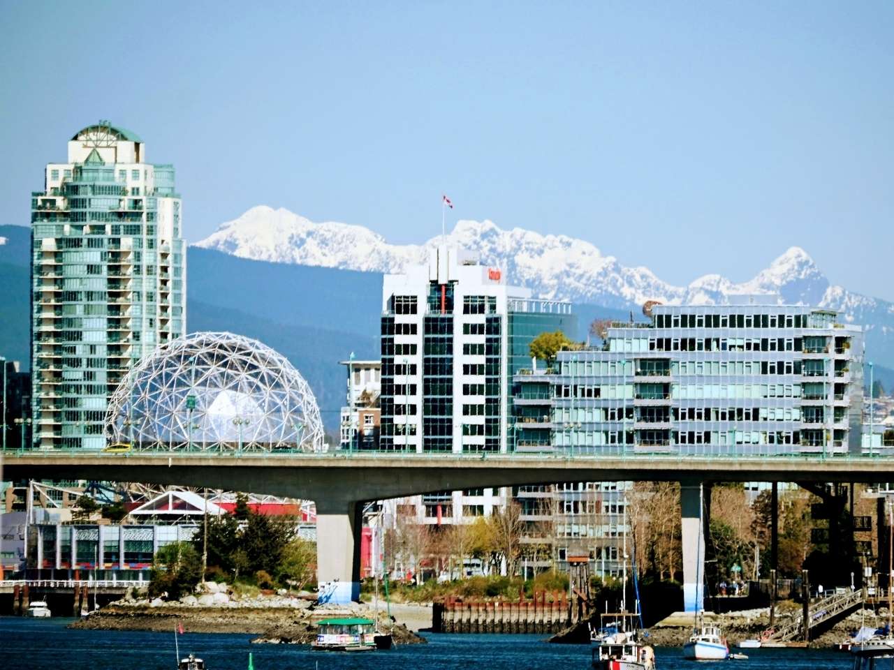 Cityscape featuring the geodesic dome of science world and modern buildings with glass facades, set against snow-capped mountains and a blue sky. Boats float in the foreground of olympic village in Vancouver