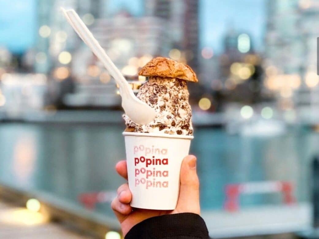 a hand holding a plastic cup with popina printed on it and filled with the famous cream puff with the city as the background