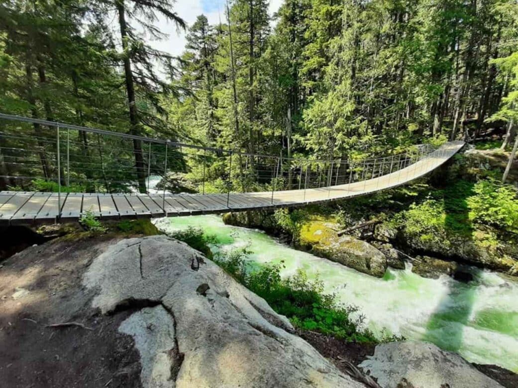 a bridge on top of a very clear lake surrounded by big trees on the way to the train wreck